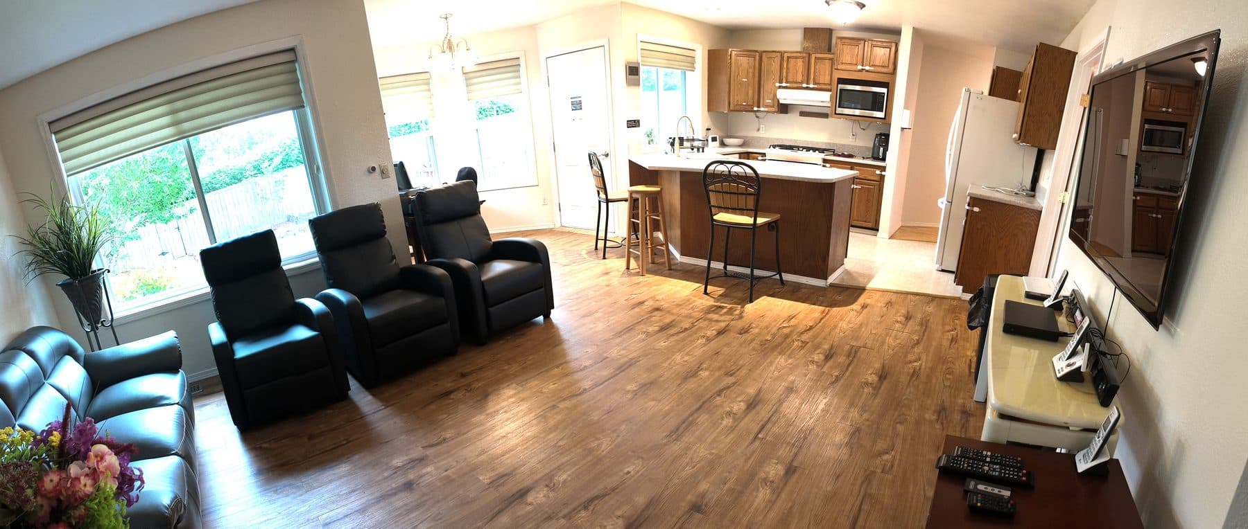 Living room with recliner seating, a wall-mounted television and a bright window, open to the kitchen